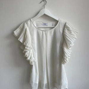 Vit blus med volangärmar Love Divine - Supersöt vit blus från Love Divine med plisserade volangärmar som ger en riktigt drömmig look. Blusen har rund halsringning och är gjord i ett lätt, flowy material som känns lyxigt. Perfekt när du vill sticka ut lite extra med dina ärmar!