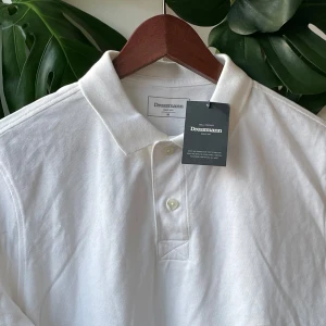 Vit pikétröja från Dressmann - Klassisk vit pikétröja från Dressmann i bomull. Tröjan har korta ärmar, krage och knäppning med två knappar framtill. Perfekt för en clean och stilren look. Passar dig som gillar enkel och tidlös stil.