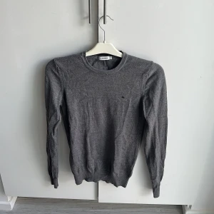 Grå stickad tröja från J.Lindeberg - Snygg grå stickad tröja från J.Lindeberg med rund halsringning och diskret logga på bröstet. Tröjan har lång ärm och ribbade muddar vid ärmslut och nederkant. Perfekt för lager-på-lager och enkel att matcha med jeans eller chinos. Köparen står för frakten!