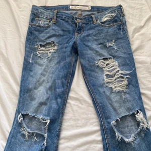 Blå slitna bootcut jeans Hollister - Blå jeans från Hollister med bootcut passform och rejält slitna detaljer på både fram- och baksida. Jeansen har klassisk femficksdesign, låg midja och är tillverkade i bomull. Dom har hål längst ner vid fötterna men inget man lägger märke till, skriv om ni vill ha fler bilder eller andra frågor!