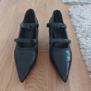 Svarta pumps med dubbla remmar - Stilrena svarta pumps i skinn med spetsig tå och låg klack. Skorna har två smala remmar över foten som ger en cool och trendig look. Perfekta för dig som gillar klassiska men edgy detaljer.