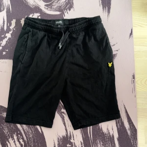 Svarta shorts från Lyle & Scott - Svarta shorts från Lyle & Scott med elastisk midja och snörning. Klassisk design med sidofickor och en bakficka. Liten gul logga broderad på ena benet. Perfekta för sommardagar och chill häng.