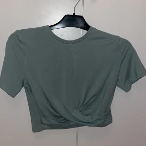 Grön topp från H&M  - Snygg croppad topp i dämpad grön färg från H&M Divided. Toppen har korta ärmar, rund halsringning och en twistad detalj framtill. Aldrig använd i fint skick 🩷