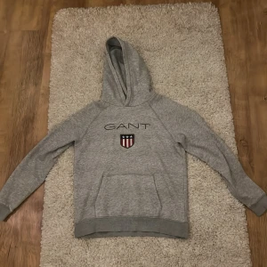 Grå hoodie från GANT - Snygg grå hoodie från GANT med klassisk logga och sköld på bröstet. Tröjan har huva, magficka och ribbade muddar. Perfekt för dig som gillar en stilren och sportig look. Passar både till jeans och mjukisbyxor.