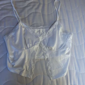 Vit satinlinnetopp med spetsdetaljer - Snygg vit linnetopp i satin med tunna axelband och vackra spetsdetaljer längs urringning och framstycke. Croppad modell som ger en trendig look, perfekt att styla med jeans eller kjol. Justerbara axelband för bästa passform.