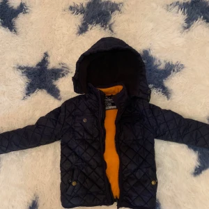 Mörkblå quiltad jacka från Tex - Quiltad mörkblå jacka från Tex med huva och orange fleecefoder. Jackan har dragkedja, tryckknappar och flera fickor framtill. Perfekt för kyliga dagar och har en snygg, sportig look med rutmönster och praktiska detaljer. Knappar till bröstfickorna har ramlat av.