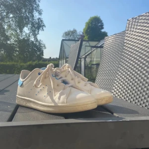 Adidas Stan Smith vita/blå sneakers - Snygga Adidas Stan Smith sneakers i vitt med blå detaljer på häl och plös. Klassisk låg modell med snörning och perforerade ränder på sidorna. Skorna har platt sula och är tillverkade i skinn och syntet. Perfekta för dig som gillar en clean och sportig look.