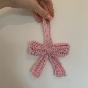 Rosa virkad rosett-accessoar - Supersöt handgjord rosett i ljusrosa. Perfekt att hänga på väskan för en gullig touch. Rosetten har en ögla upptill för enkel fastsättning och är mjuk och fluffig. Rosetten är 11cm bred och 9cm hög