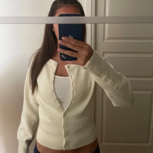 Krämvit kofta med knappar - Superfin krämvit kofta med rund hals och långa ärmar. Koftan har ribbade muddar och många små knappar framtill samt på ärmsluten. Den är stickad och har en croppad passform som sitter snyggt i midjan. Perfekt att styla över ett linne eller t-shirt. Storleken är S, dock passar den dig som har Xs också. Vill även tillägga att den aldrig använts, då jag tycker att den är superfin men lite kort eftersom jag har en lång öcerkropp… Pris kan alltid diskuteras! 