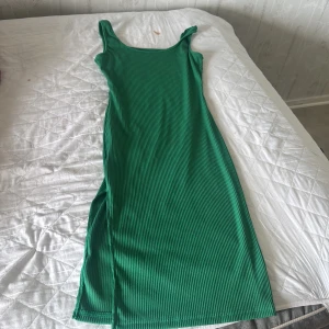 Grön ribbad långklänning - Snygg grön långklänning med ribbad struktur och ärmlös design. Klänningen har en slits på sidan och smala axelband, perfekt för varma dagar. Enkel och stilren modell som är lätt att matcha med accessoarer.