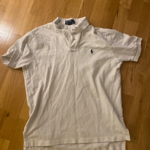 Vit pikétröja från Polo Ralph Lauren - Vit pikétröja från Polo Ralph Lauren med klassisk krage och knappar framtill. Ikoniska broderade loggan på bröstet. Tillverkad i 100% bomull, perfekt för en clean och stilren look.