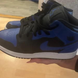 Nike Air Jordan 1 blå och svarta - Säljer ett par Nike Air Jordan 1 i blått och svart skinn med vita sulor. Klassisk high-top siluett med svart snörning och blå detaljer på både sidor och häl. Perfekta för dig som vill sticka ut med en ikonisk sneaker.