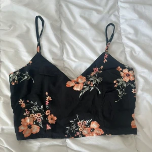 Svart blommig croppad linnetopp - Supersöt svart croppad linnetopp med orange och rosa blommor. Justerbara smala axelband och smockad rygg med en stor rosett som detalj. Toppen har v-ringning och är perfekt för varma dagar. Lätt och luftig känsla, passar till både jeans och kjol