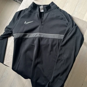 Svart Nike half zip träningströja - Svart långärmad träningströja från Nike med vit logga på bröstet och en snygg grå rand över bröstet. Tröjan har half zip och är gjord i lätt polyester med Dri-FIT-teknik som andas. Perfekt för träning eller chill dagar. Skick 10/10, storlek Large.