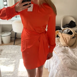 Orange kort klänning från Vila - Superfin orange kort klänning från Vila med rund hals, långa ärmar som man kan knyta i midjan.Klänningen har en snygg detalj med liten öppning i ryggen och är tillverkad i ett lätt, slätt material som ger ett stilrent intryck.