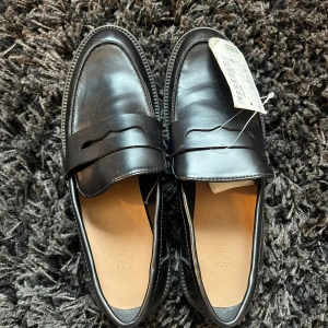 Svarta loafers från H&M i skinnimitation - Stilrena svarta loafers från H&M i skinnimitation med klassisk pennyloafers-design och rund tå. Perfekta för en clean och dressad look. Helt nya och aldrig använda hann bara inte lämna tillbaka: