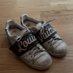Louis Vuitton vita sneakers med logga - Snygga vita sneakers från Louis Vuitton med breda svarta remmar över vristen där det står 'Louis Vuitton' i vit text. Skorna har snörning, rund tå och detaljer i skinn. Perfekta för dig som vill sticka ut med en lyxig streetstyle. Ny pris 7000kr