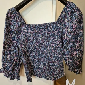 Blommig smockad blus med puffärm - Supersöt blommig blus med smockad kropp och puffiga långa ärmar. Färgerna går i blått, lila och rosa med små vita blommor. Fyrkantig ringning och volangdetaljer vid ärmslut och nederkant. Perfekt för dig som gillar romantisk stil!