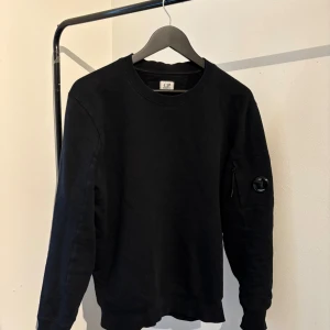 Svart sweatshirt från C.P. Company - Svart sweatshirt från C.P. Company med rund hals och lång ärm. Tröjan har en ikonisk linsdetalj och dragkedjeficka på ena ärmen. Tillverkad i mjuk bomull. Köpt för ca 5 månader sedan och har använt ett fåtals gånger. Inga skador eller slitningar, bara någon repa på linddetaljen, men inget märkvärdigt.