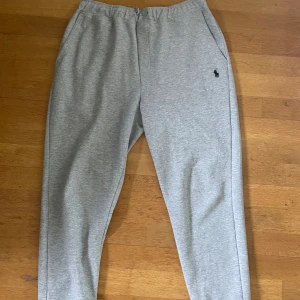 Grå mjukisbyxor från Polo Ralph Lauren - Snygga ljusgrå mjukisbyxor från Polo Ralph Lauren med klassisk broderad logga på ena benet. Byxorna har resår i midjan och sidofickor, tillverkade i mjuk bomullsmix för maximal komfort. Perfekta för chill dagar eller när du vill vara bekväm.