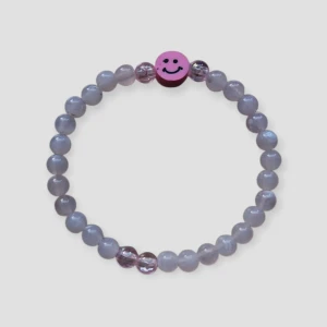 Armband med rosa pärlor och smiley - Stretchigt armband med ljusrosa pärlor och detaljer i transparent rosa. Mitt på sitter en rosa pärla med svart smiley-ansikte som ger en lekfull touch. (Den ser lila ut på bilden men är rosa)
