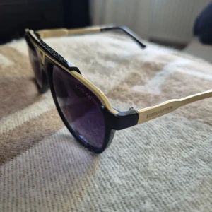 svarta och guldiga solglasögon - Snygga solglasögon från med svart båge och guldiga detaljer. ikonisk V-detalj framtill. Klassisk fyrkantig form med exklusiv känsla och logotyp på skalmarna. Perfekta för dig som vill sticka ut med lyxig stil.