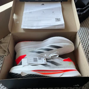 Adidas Adizero SL2 vita löparskor - Säljer ett par Adidas Adizero SL2 sneakers i vitt med svarta detaljer och orange inslag. Skorna har en sportig, aerodynamisk form och är tillverkade i lätt syntetmaterial med snörning och platt sula. Perfekta för dig som gillar att springa snabbt och stilrent. Helt nya med prislapp och orginal förpackning 