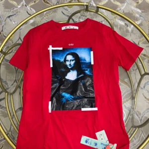 Röd Off-White t-shirt med Mona Lisa print - Röd t-shirt från Off-White med ikoniskt Mona Lisa-tryck i blått och svart framtill och stora vita pilar på ryggen. Klassisk passform, rund hals och korta ärmar. Materialet är mjuk bomull och t-shirten har tydliga streetwear-vibbar.