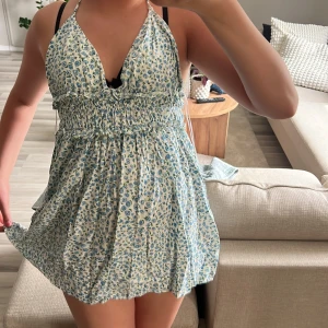 Blommig kortklänning från Zara XS - Supersöt kortklänning från Zara i vitt med blått blommönster. Halterneck med knyt i nacken, smockad midja och luftig kjol. Perfekt för varma dagar och ger en fräsch sommarkänsla. Materialet är lätt och känns skönt mot huden. Den är i ett nytt skick med inga skador.JAG STÅR INTE FÖR NÅGON FRACKT!