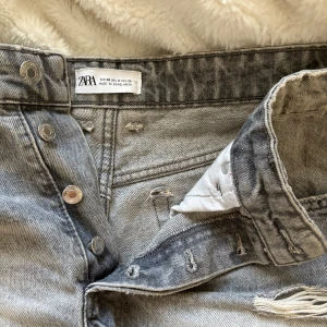 Grå jeansshorts från ZARA - Snygga grå jeansshorts från ZARA med slitna detaljer och fransig nederkant. Klassisk femficksmodell med hög midja och knappar framtill. Perfekta för en chill sommarlook.