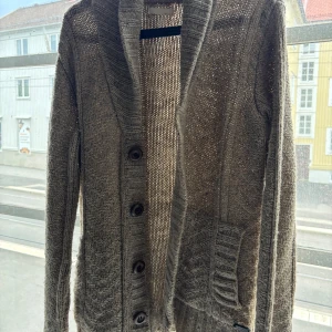 Stickad cardigan från suit - Snygg beige stickad kofta med stora knappar framtill och två fickor. Koftan har lång ärm, bred krage och ett mysigt, grovstickat mönster. Perfekt att slänga på sig när det är kyligt ute.