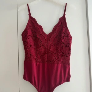 Vinröd spetsbody med smala axelband - Snygg vinröd body med blommig spets upptill och slät nederdel. Djup v-ringning och smala, justerbara axelband. Perfekt för att lyfta vilken outfit som helst med sin detaljerade spets och feminina look