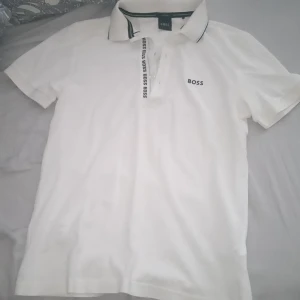 Vit piké t-shirt från BOSS mötts upp i gbg - Vit piké t-shirt från BOSS med klassisk krage och korta ärmar. Diskret BOSS-logga på bröstet och tryckt logga längs knäppningen. Snygga svarta detaljer på kragen. Perfekt för en clean och stilren look.