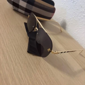 Burberry solglasögon med guldiga bågar - Snygga Burberry solglasögon med tunna guldiga metallbågar och mörka linser. Sidorna har den klassiska Burberry-randiga detaljen. Kommer med ett fodral i beige, svart och brunt rutigt mönster. Perfekt accessoar för att lyfta din stil.