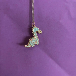 Halsband med blå dinosaurie-hänge - Sött halsband med ett hänge i form av en blå dinosaurie med guldiga detaljer och små rosa inslag. Dinosaurien håller en liten blomma i munnen. Kedjan är tunn och silverfärgad, perfekt för dig som gillar unika och lekfulla smycken.