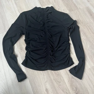Svart långärmad topp från Gina Tricot - Svart långärmad topp från Gina Tricot med snygg rynkad detalj längs hela mitten fram- och baktill. Topp i stretchigt material som sitter tajt och ger en clean look. Perfekt att styla med jeans eller kjol för en trendig vibe. 