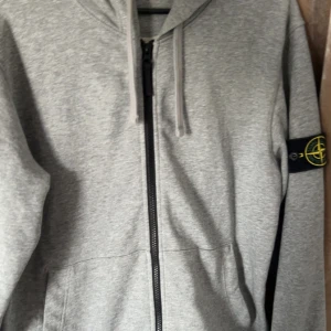Grå zip-hoodie från Stone Island - Grå hoodiejacka från Stone Island med dragkedja framtill och klassisk logga på ärmen. Jackan har huva med snörning och två fickor framtill. Perfekt för en chill och stilren look.