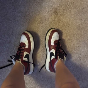 Nike Air Force 1 vinröd/vit sneakers - Snygga Nike Air Force 1 sneakers i vinrött och vitt med svarta detaljer och svart swoosh. Klassisk låg modell med svart snörning, perforerad tå och robust sula. Ovandel i skinn och textilfoder för extra komfort. Perfekta för dig som vill sticka ut med stil.