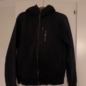 Svart hoodiejacka från Sail Racing M - Svart hoodiejacka från Sail Racing med dragkedja hela vägen och bröstficka med logga. Jackan har huva, långa ärmar och är tillverkad i mjukt material som känns skönt mot kroppen. Perfekt för dig som gillar en clean och sportig stil.