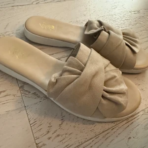 Beige sandaler med rosett från So All - Snygga beige sandaler från So All med breda band i mockaliknande material och stor rosett framtill. Platt sula och öppen tå gör dem perfekta för varma dagar. Stilren och trendig design som passar till många outfits.