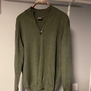 Grön half zip tröja Oscar Jacobson - Mörkgrön stickad tröja från Oscar Jacobson i storlek L men passar M. Tröjan har en ribbad krage och en snygg half zip-dragkedja framtill. Tillverkad i Italien och passar perfekt till lager-på-lager-stil under höst och vinter.