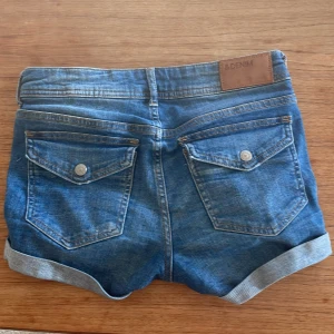 Blå jeansshorts med fina fickor  från H&M, strl 34 - Snygga blå jeansshorts från H&M i klassisk modell med regular waist. Shortsen har uppvikt benskör,med knappar på bakfickorna. Perfekta för sommardagar och enkel att matcha med allt. Shortsen är i storlek 34 men passar mig som har 32-34! 