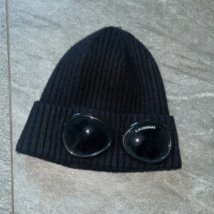 Svart mössa från C.P. Company - Svart ribbstickad mössa från C.P. Company med ikoniska goggle-linser framtill. Mössan är tillverkad i mjuk ull och har en uppvikt kant där linserna sitter fast. Perfekt för dig som vill sticka ut med streetwear-vibe.