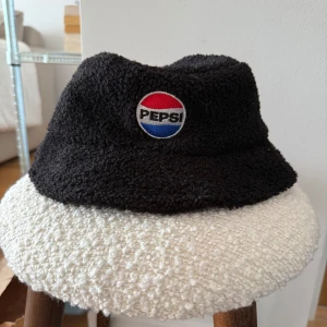 Svart Pepsi bucket hat i teddy - Svart bucket hat i mjuk teddy med klassisk Pepsi-logga framtill i rött, blått och vitt. Unisex storlek och jätteskön. Superfint skick.