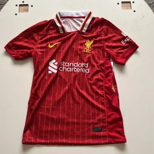 Liverpool FC röd matchtröja Nike size men small - Liverpool FCs officiella matchtröja i rött med gula detaljer och tunna gula ränder. Broderat klubbmärke på bröstet, Nike-logga och sponsortryck. Klassisk krage med vita och gula inslag. Tillverkad i lätt och ventilerande polyester.