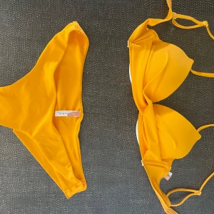 Gul/orange bikini Nelly - Snygg gul bikini från Nelly med vadderad trekants-bh och justerbara axelband. Storlek SMALL. Underdelen har klassisk normal midja och enkel, stilren look. Perfekt för stranden eller poolen, färgen poppar verkligen i solen! 
