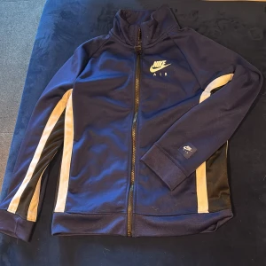 Nike Air blå träningsjacka M ca 146 - Nike Air vindjacka i mörkblått med vita och svarta ränder längs ärmarna. Jackan har hög krage, hel dragkedja och stora Nike Air-loggor på bröstet och ryggen. Tillverkad i polyester, perfekt för en sportig look.