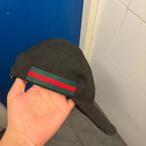 Svart Gucci keps med monogram - Svart keps från Gucci med diskret GG-monogram över hela tyget och klassiska gröna och röda ränder på sidorna. Justerbar rem baktill med metallspänne. Snygg och stilren accessoar som lyfter din outfit.
