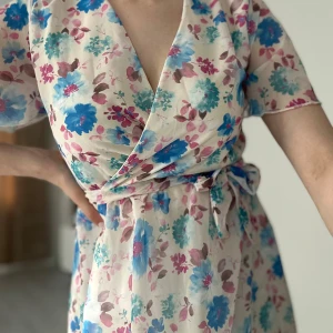 Blommig omlottklänning med knytband - Superfin långklänning med blommigt mönster i blått, rosa och turkost på ljus botten. Omlottmodell med v-ringning, korta ärmar och knytband i midjan. Perfekt för sommarens alla tillfällen!