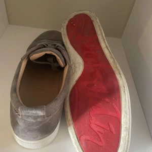 Grå loubs utan spikar - Snygga grå sneakers i mocka från Louboutin med den ikoniska röda sulan. Skorna har snörning, rund tå och vit platt sula. Perfekta för dig som vill ha en stilren men ändå lyxig look.